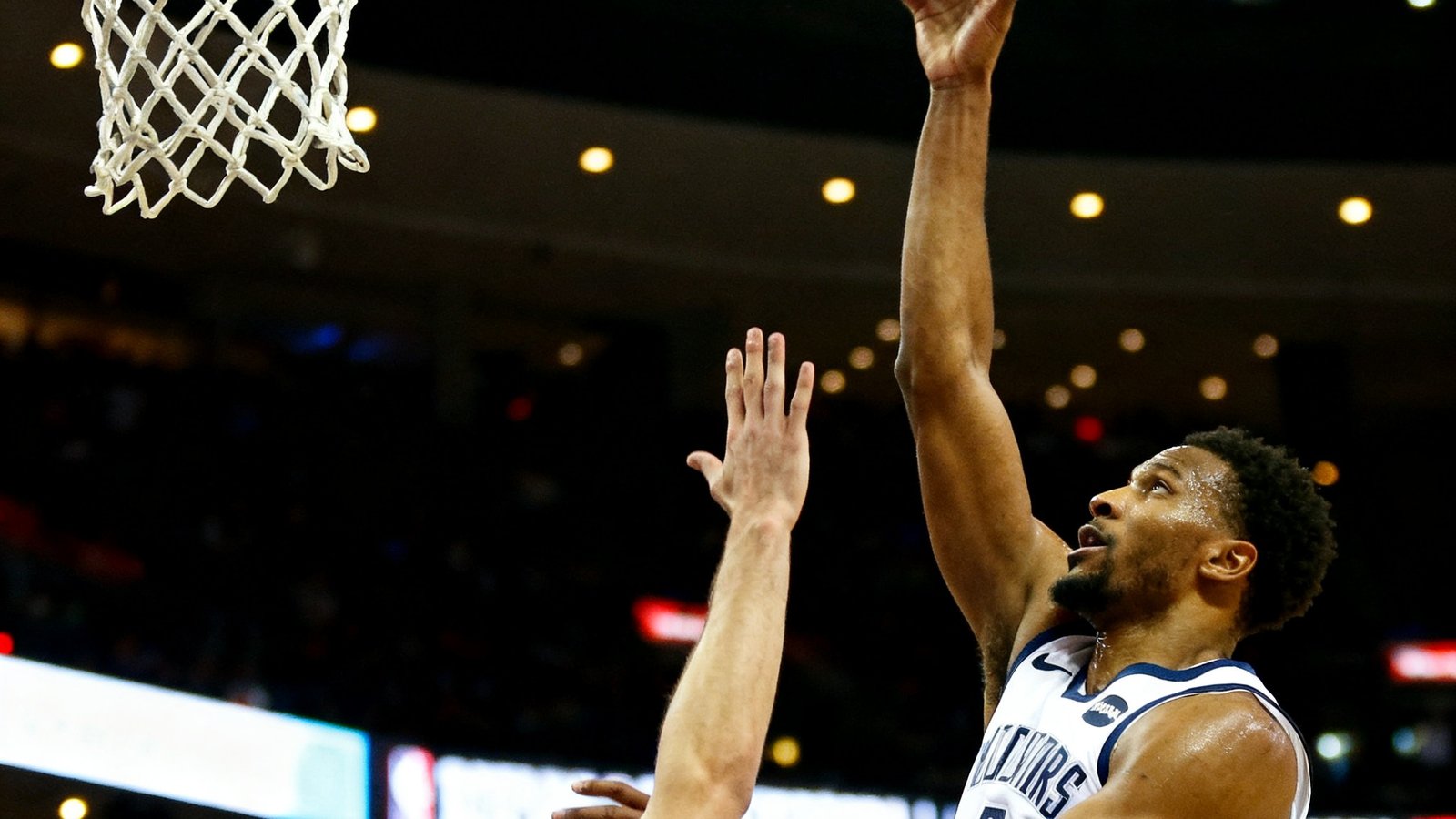 NBA常规赛比赛现场，一名身穿白色球衣的球员在三分线外跳投，篮球划出弧线飞向篮筐，防守球员高举手臂试图干扰